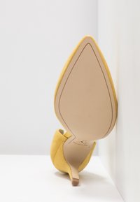 Chaussure à talon haut en daim jaune à bout pointu, reposant sur son talon contre un mur et une surface blancs, montrant la semelle beige lisse.