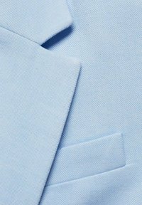 Détail en gros plan du col de veste en tissu texturé bleu clair et de la poche à rabat.