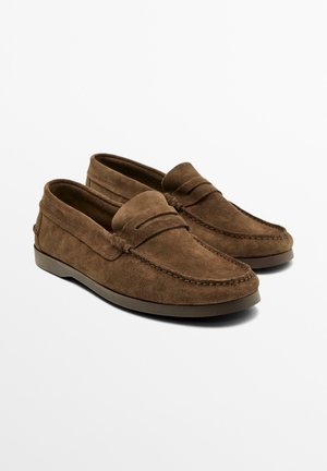 Bruine suède leren loafers met gestikte details, mocassinstijl neus en lage platte rubberen zool, afgebeeld tegen een effen witte achtergrond.