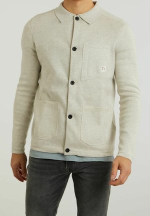 Mężczyzna w jasnobeżowym zapinanym na guziki kardiganie z trzema kieszeniami na niebieskiej koszuli i w ciemnoszarych dżinsach, stojący na jednolitym tle.