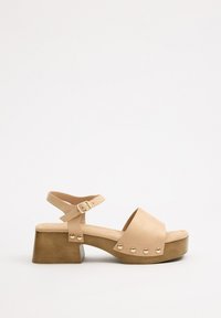 Sandalia de plataforma beige con parte superior de cuero suave, tira ancha, hebilla en el tobillo y tachuelas redondas a lo largo de la base de madera. Diseño de suela plana.