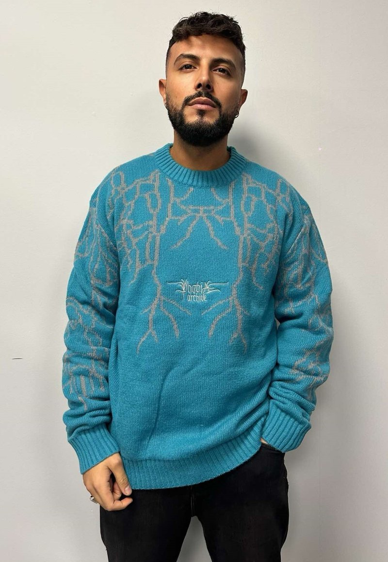 Maglione turchese a maglia con un design testurizzato e dettagli beige. Presenta polsini e orlo a costine. Sfondo semplice e di colore chiaro.