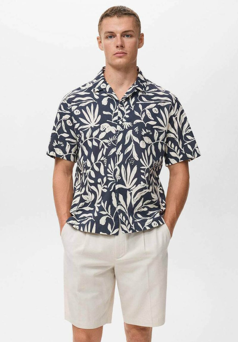 Camisa de manga corta azul marino con patrón de hojas blancas, hecha de tela ligera. Combinada con pantalones cortos de color claro, el modelo posa frente a un fondo blanco.