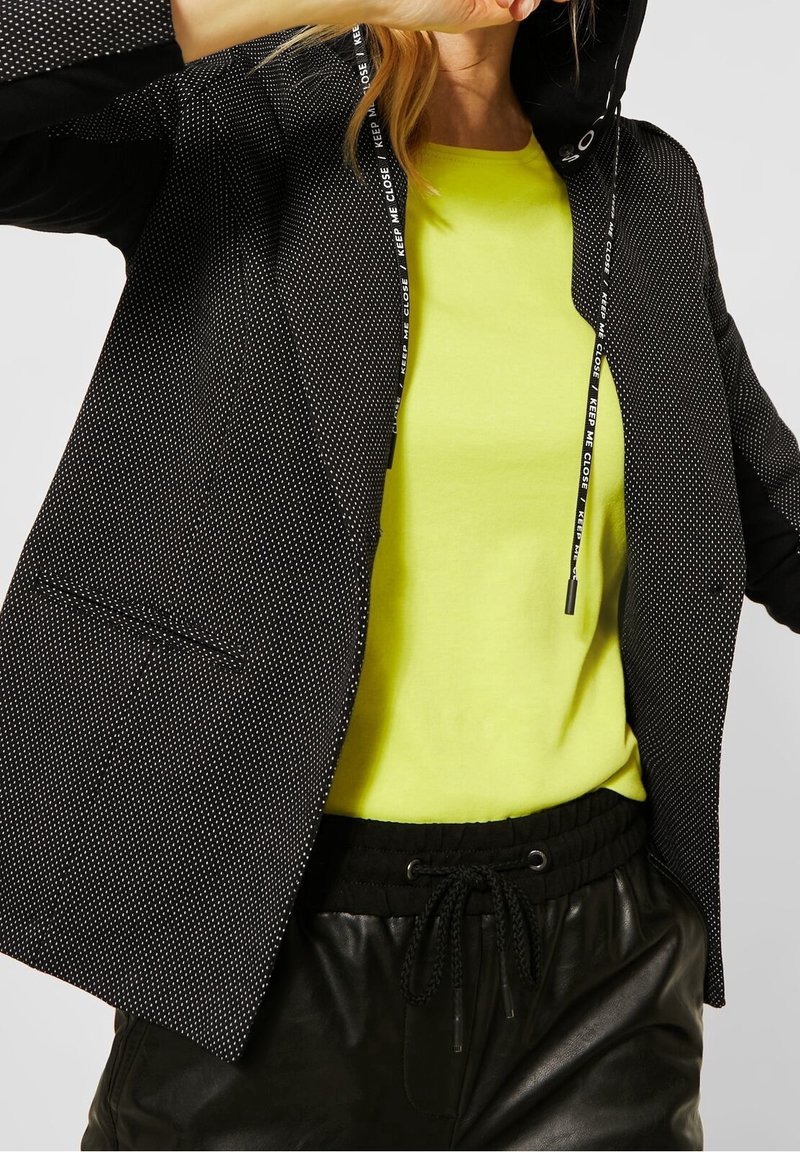 Personne portant un haut jaune fluo, un blazer noir à pois avec des cordons portant l'inscription "GARDES-MOI PROCHE", et un pantalon en cuir noir avec cordon de serrage.