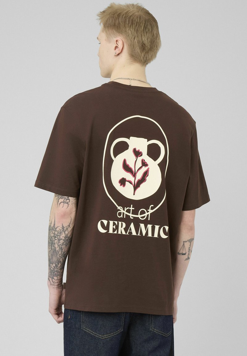 Jeune personne aux cheveux blonds portant un T-shirt marron avec un motif de vase en céramique et de fleurs, accompagné du texte « art of CERAMIC » au dos.
