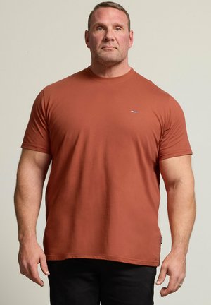 Muskeløs mand, der står med fronten mod kameraet iført en ensfarvet rustfarvet kortærmet skjorte og sorte bukser mod en neutral baggrund.