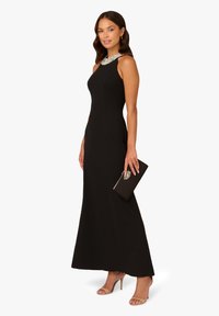 Schwarzes Abendkleid mit taillierter Silhouette, asymmetrischem Ausschnitt und mit Perlen verziertem Kragen. Kombiniert mit einer schwarzen Clutch und nudefarbenen High Heels.