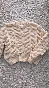 Cardigan beige lavorato a maglia con un motivo a onde testurizzate, orlo e polsini a coste, e bottoni a forma di fiore. Materiale morbido, stile cropped.