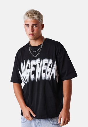 Joven con el pelo corto y decolorado lleva una camiseta negra de gran tamaño con texto blanco distorsionado y collares de doble cadena, posando contra un fondo blanco.