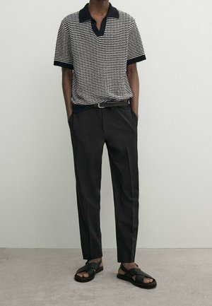 Uomo che indossa una polo a maniche corte in maglia bianca e nera, cintura nera, pantaloni su misura neri e sandali neri aperti, in piedi su pavimento grigio.