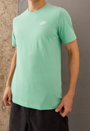 Mężczyzna w miętowo zielonej koszulce Nike z białym logo i ciemnych spodniach na tle teksturowanych kamiennych i szarych ścian.