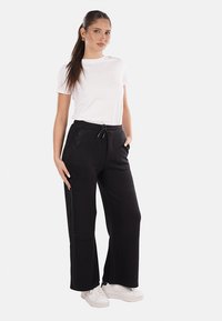 Pantaloni sportivi neri con gamba larga, vita con cordoncino e dettagli decorativi in strass sulle tasche, abbinati a una maglietta bianca semplice e a sneakers bianche.