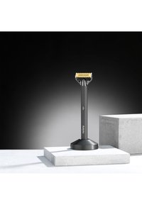 Svart elektrisk barberhøvel med et rektangulært gullblad, montert på et matt svart stativ. Har et grep og en på/av-knapp.