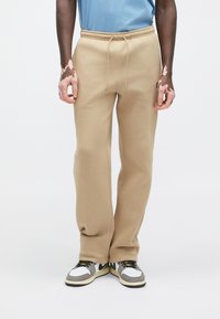 Bézs színű melegítőnadrág, rugalmas derékpánttal és húzózsinóros részlettel. Az anyag puha és rugalmas, színes sportcipőkkel kombinálva.