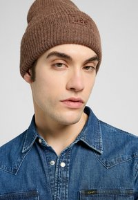 Beanie a coste marrone con dettagli del logo. Indossato con una camicia di denim blu con bottoni a pressione e taschino sul petto.