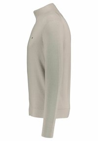 Pull léger beige avec un col montant, texture côtelée et manches longues. Présente des panneaux latéraux de couleur vert clair contrastants et un logo minimal.