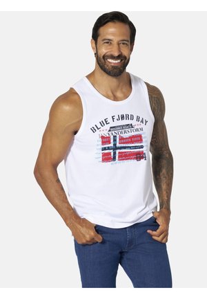 Man with beard and tattooed arm wearing white sleeveless shirt with "Blue Fjord Bay" text and red, white, and blue graphic, smiling with hands in pockets.