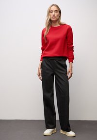 Roter Rundhals-Sweatshirt, schwarze weite Beine-Hosen, weiße Sneakers mit beigen Sohlen auf einem grauen Boden vor einer hellen Wand.