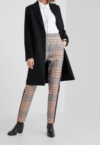 Long manteau noir sur une chemise blanche avec des bordures noires, associé à un pantalon à carreaux gris avec des lignes orange et bleues, et des bottines noires à la cheville.