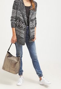 Grå, texturerad kofta med geometriska mönster, bärs över en svart topp; kombinerad med ljusblå jeans och en metallisk silverhandväska.