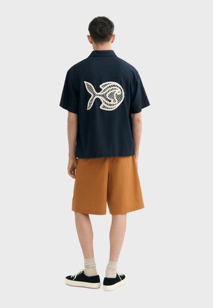 Homme portant une chemise noire à manches courtes avec un motif de poisson blanc dans le dos, un short marron, des chaussettes beiges et des baskets noires, vu de dos.