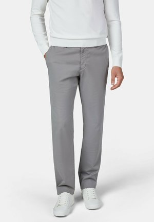 Man wearing grey straight-leg pants, white long-sleeve top, and white sneakers standing with one hand in pocket.