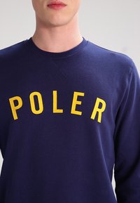 Sweat-shirt bleu marine à col ras du cou avec lettrage jaune "POLER". Tissu doux, encolure côtelée, et coupe décontractée pour le confort et une grande liberté de mouvement.