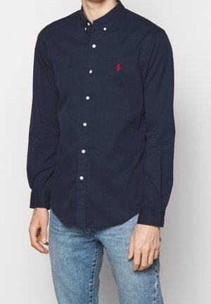 Mies pukeutunut tummansiniseen, napitettuun pitkähihaseen paitaan, jossa on punainen logo rinnassa, ja vaaleansinisiin farkkuihin, seisoo yksiväristä taustaa vasten.