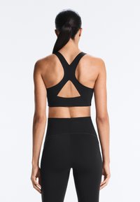 Sötét haját lófarokba kötött nő, fekete, versenyhátsó sportmelltartót és magas derekú fekete leggingset visel, hátulnézetből.