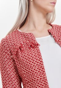 Rode en witte getextureerde jas met een geruit patroon, voorzien van een rimpelachtige kraag. Draagt over een witte top, die een getailleerd ontwerp toont.