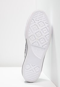 Semelle d'une basket Converse All Star noire et blanche montrant un motif de bande de roulement détaillé et le logo, inclinée contre un mur blanc.