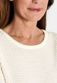 Frau, die sanft lächelt, trägt einen weißen, strukturierten Strickpullover, mit sichtbarem Hals- und Schulterbereich vor einem schlichten Hintergrund.