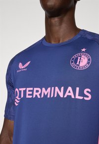 T-shirt de sport bleu à manches courtes, avec un col rond, un logo de sponsor rose et un emblème d'équipe, en tissu texturé et avec des accents à motifs sur les épaules.