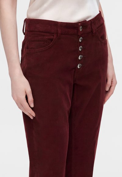 Femme portant un pantalon en velours côtelé rouge foncé taille haute avec cinq boutons argentés et des poches, les mains reposant le long du corps.