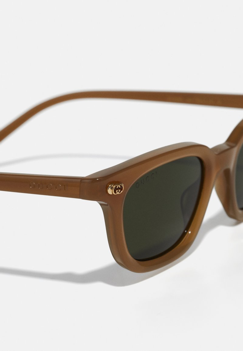 Occhiali da sole in plastica marrone con lenti verde scuro, dotati di una montatura quadrata e un dettaglio del logo dorato sul lato. Texture liscia e design robusto.