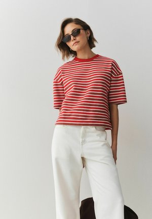 Frau mit schwarzen Sonnenbrillen, rot-weiß gestreiftem kurzärmligem Pullover und hoch taillierter weißer Hose steht vor einer einfarbigen hellen Wand.