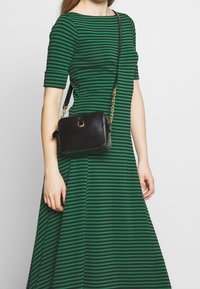 Kvinna klädd i en grön och svart randig klänning med ärmar som når till armbågen, bär en svart läder crossbody-väska med ett guld kedjesläp.