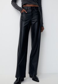 Pantalon large en similicuir noir à texture lisse, taille haute, poches avant et détails de surpiqûres contrastées.