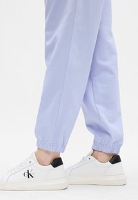 Heledad lilla dressipüksid elastsete pahkluude juures, koos valgete tossudega, millel on must konts ja küljes "CK" logo.