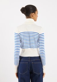 Strikket turtleneck-genser i hvit med blå horisontale striper, rillemønster og tettsittende ermer, kombinert med mørke høyhalsede jeans.