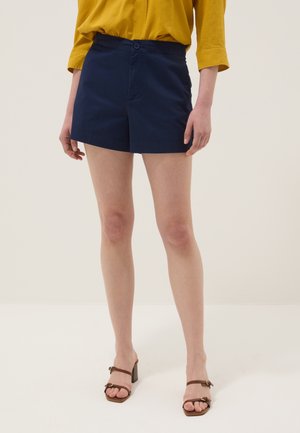 Personne portant un short bleu marine sur mesure, un haut jaune à manches longues et des sandales marron à talons, debout sur un fond clair uni.