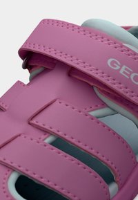 Ροζ παιδικό σανδάλι με ρυθμιζόμενους ιμάντες Velcro και ορατή ραφή, με μερικώς ορατό όνομα επωνυμίας στον πάνω ιμάντα.