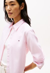 Camicia a maniche lunghe a righe rosa chiaro, realizzata in cotone, con colletto classico e un piccolo logo sul lato sinistro del petto. Maniche arrotolate visibili.