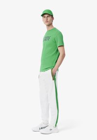 Grünes T-Shirt mit kurzen Ärmeln und Logo, weißer Jogginghose mit grünen Seitenstreifen und weißen Sneakers. Das Modell trägt eine passende grüne Cap.