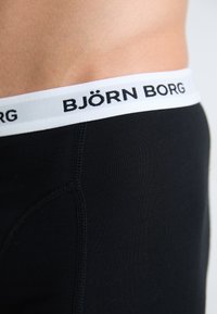 Sorte boxer shorts laget av glatt stoff, med en bred hvit midjebånd med svart "BJÖRN BORG"-skrift og kontrasterende sømmer.
