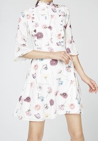 Blommig klänning i vitt tyg med korta utställda ärmar, prydd med pastellfärgade blommor i rosa, lila och blått, med hög krage och veckdetaljer.