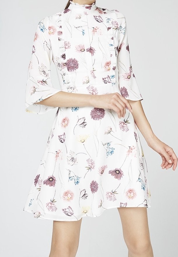 Blommig klänning i vitt tyg med korta utställda ärmar, prydd med pastellfärgade blommor i rosa, lila och blått, med hög krage och veckdetaljer.
