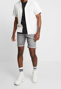 Camisa blanca de manga corta, camiseta negra debajo, pantalones cortos de mezclilla gris con dobladillos enrollados, zapatillas blancas y calcetines rayados hasta el tobillo.
