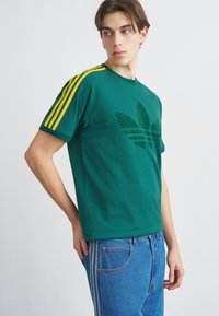 Jauns vīrietis zaļā T-kreklā ar dzeltenām svītrām uz piedurknēm un lielu logotipu, tērpies zilās džinsās, skatās sāniski pret vienkāršu fonu.