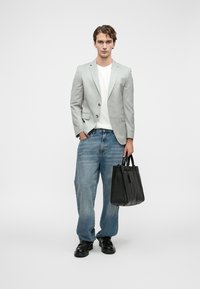 Νεαρός άνδρας wearing ελαφρύ γκρίζο σακάκι, λευκό πουκάμισο, φαρδιά μπλε τζιν, μαύρα παπούτσια, κρατώντας μαύρη δερμάτινη τσάντα, στέκεται μπροστά σε λευκό φόντο.